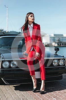 Attractive girl standing next to a retro sport car on the sun. Fashion woman in a red suit and sunglasses waiting near classic car