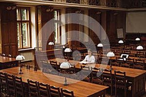 Attractive girl student in a white blouse sitting alone in an atmospheric public library with a book and looking at the camera