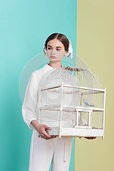 attractive girl in trendy white outfit with parrot in cage on turquoise