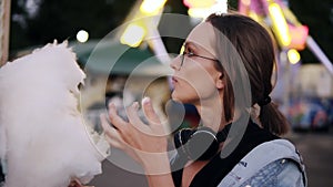 An attractive girl walking in the amusment park, eats white cotton candy. Girl wearing casual clothes, headphones on her