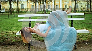 Attractive girl in white and blue dress lies on bench with legs in high heeled shoes riased on back of bench in parkway