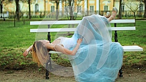 Attractive girl in white and blue dress lies on bench with legs in high heeled shoes riased on back of bench in parkway