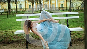 Attractive girl in white and blue dress lies on bench with legs in high heeled shoes riased on back of bench, she takes
