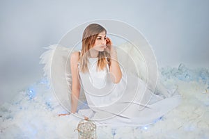 An attractive girl in a white tunic with large white wings behind her back poses while sitting in white luminous clouds