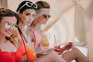 attractive girlfriends in swimwear and sunglasses drinking cocktails in bungalow