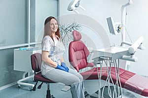 Attractive good-looking portrait of female dentist in clinic sitting on chair near dental unit with hands in blue gloves, smiling