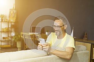 Attractive grey-haired man relaxing on a couch at home, using mobile phone. Handsome man in glasses texting in cellura phone