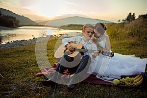 Attractive groom plays the guitar and his charming bride enjoys it during their picnic on the river bank during the