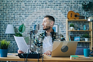 Attractive guy recording podcast using microphone holding piece of paper indoors