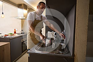 Attractive handsome Caucasian young man in beige chefs apron, filling a stainless steel saucepan with running water