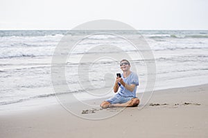 Attractive and handsome man on his 30s sitting on the sand relaxed on the beach laughing in front of the sea texting on mobile pho