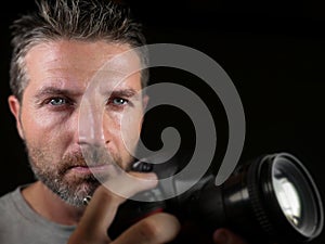 Attractive and handsome man on his 30d holding professional reflex photo camera next to his face isolated on black background in