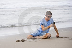 Attractive and handsome man on his 30s sitting on the sand relaxed on the beach laughing in front of the sea texting on mobile pho