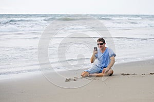 Attractive and handsome man on his 30s sitting on the sand relaxed on the beach laughing in front of the sea texting on mobile pho