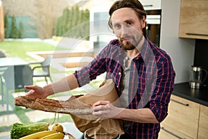 Attractive handsome young Caucasian man unpacking the eco paper bag with healthy food and showing the whole grain bread or freshly