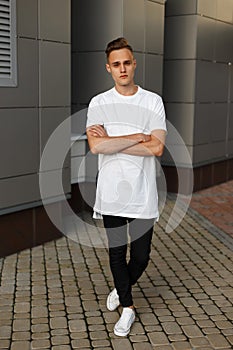 Attractive handsome young man in a white stylish T-shirt in black jeans with a clock is standing on the street