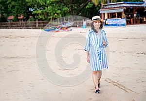 Attractive happy Asia woman walking alone on the island beach at Thailand. Wearing dress and hat, holding sun glasses and looking