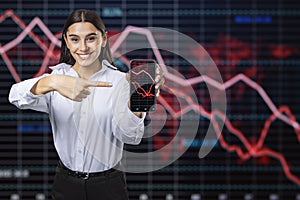 Attractive happy caucasian businesswoman pointing at cellphone with glowing downward red forex chart on blurry background.