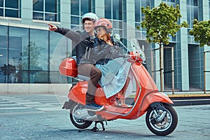 Attractive happy couple, a handsome man and female riding together on a red retro scooter in a city.