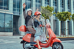 Attractive happy couple, a handsome man and female riding together on a red retro scooter in a city.