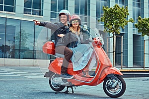 Attractive happy couple, a handsome man and female riding together on a red retro scooter in a city.