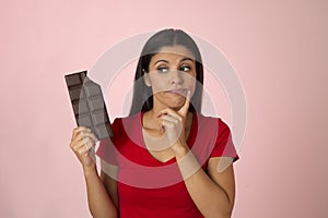 Attractive and happy hispanic woman in red top thinking and doubting holding chocolate bar on pink background