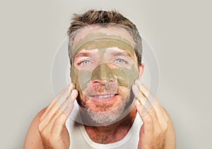 Attractive and happy man looking to himself in bathroom mirror with green cream on his face applying facial mask skin care product