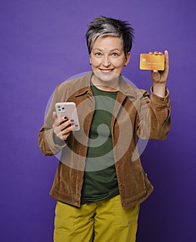 Attractive happy middle-age woman holding credit card, using cell phone for online shopping or payments isolated in studio. Online