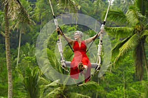 Attractive happy middle aged 40s or 50s Asian Indonesian woman with grey hair riding rainforest swing carefree swinging and