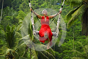 Attractive happy middle aged 40s or 50s Asian Indonesian woman with grey hair riding rainforest swing carefree swinging and