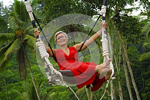 Attractive happy middle aged 40s or 50s Asian Indonesian woman with grey hair riding rainforest swing carefree swinging and