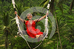 Attractive happy middle aged 40s or 50s Asian Indonesian woman with grey hair riding rainforest swing carefree swinging and