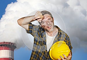 Attractive happy and tired contractor or construction worker man holding safety helmet and dirt stained face at smoking factory