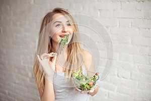 Attractive happy woman holding bowl of fresh vegetable salad in hands eating with the fork, enjoing healthy food