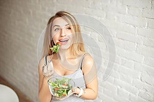 Attractive happy woman holding bowl of fresh vegetable salad in hands eating with the fork, enjoing healthy food