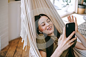 Attractive happy woman lying at the hammock, speaking on video chat, waving
