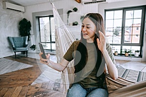 Attractive happy woman sitting at the hammock, speaking on video chat, waving