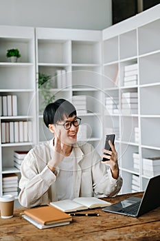 Attractive happy young Asian student studying at the college library, sitting at the desk, using a laptop, tablet and headphones