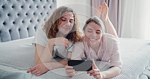 Attractive happy young couple laying on bed in the bedroom, making silly faces, taking a selfie using mobile