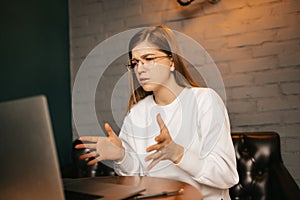 Attractive happy young girl sitting at the table, using laptop computer, video chatting, waving her arms and pointing