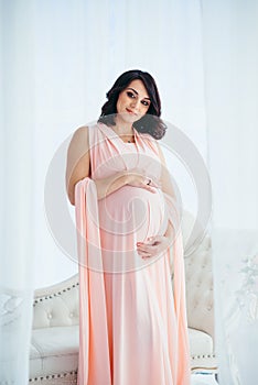 The attractive happy young pregnant woman in light pink dress stroking her belly in the white modern studio.