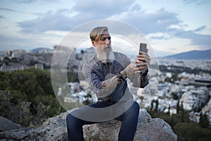 Attractive hipster with a beard is sitting on a rock high over Athens City with Acropolis view and makes a selfie