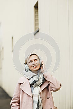 Attractive hipster blonde girl in a pink coat walks around beautiful authentic old town and listening to music on