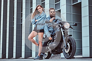 Attractive hipster couple - bearded brutal male in sunglasses and jeans jacket sitting on a retro motorcycle and his