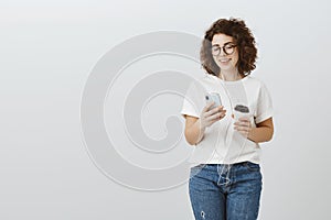 Attractive hipster girl cannot imagine life without smartphone and coffee. Carefree successful curly-haired woman in