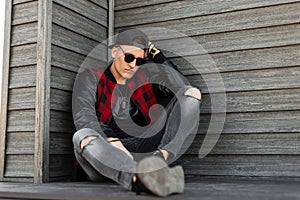 Attractive hipster young man in trendy dark sunglasses in a stylish red and black checkered jacket with leather sleeves in jeans