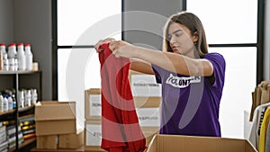 Attractive hispanic woman in casual t-shirt volunteering at charity center, elatedly packing clothes donations in a cardboard box