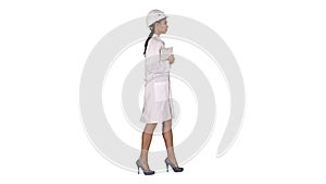 Attractive Hispanic woman in white lab coat and white safety hard hat walking holding notebook or tablet on white