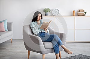 Attractive Indian lady with cup of aromatic coffee and open book relaxing in armchair at home