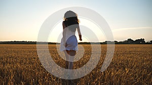 Attractive joyful woman in white dress running through field of wheat at sunset. Follow to young carefree girl enjoying